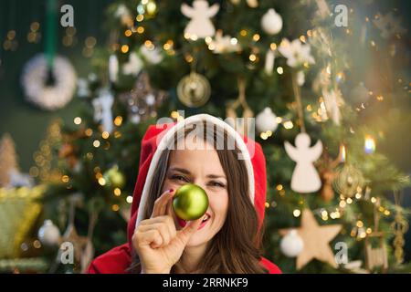 Natale. Donna elegante sorridente con cappello di Babbo Natale e palla di Natale vicino all'albero di Natale nella casa moderna. Foto Stock