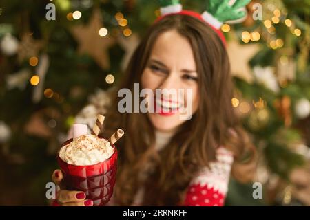 Natale. Primo piano su una casalinga moderna e felice di mezza età con un cocktail di cioccolata calda festoso vicino all'albero di Natale nel moderno soggiorno. Foto Stock