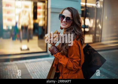 Ciao november. felice donna moderna di 40 anni con trench arancione con borse della spesa e foglie gialle autunnali vicino al negozio in città. Foto Stock