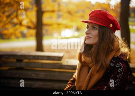 Ciao autunno. penosa donna di tendenza di 40 anni con cappello rosso con sciarpa seduta sulla panchina nel parco cittadino. Foto Stock