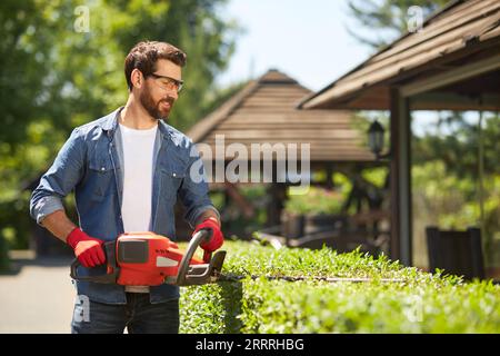 Giardiniere di mezza età che utilizza tagliasiepi elettrici per tagliare siepi nelle giornate di sole. Vista laterale dell'uomo dai capelli scuri che forma un arbusto di bosso coltivato sul cortile. Concetto di attrezzatura da giardinaggio. Foto Stock