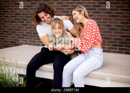 Famiglia con una madre, un padre e una figlia seduti fuori sui gradini di una veranda anteriore di una casa in mattoni Foto Stock