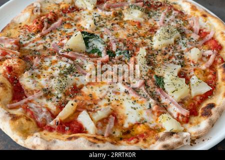 Primo piano di una pizza intera con crosta sottile di ananas e prosciutto su un piatto abbondante di deliziosi ingredienti e cosparsa di origano secco saporito Foto Stock
