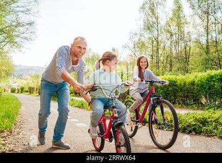 Sorridente padre con due figlie durante una passeggiata all'aperto. Insegna alla ragazza più giovane a fare una bicicletta. Amano stare insieme nel parco estivo della città. Ha Foto Stock