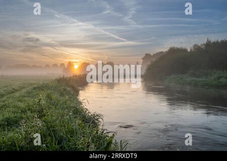 Una dolce scena fluviale con flusso costante e sponde erbose con nebbia e sole che si innalza sul lontano orizzonte Foto Stock