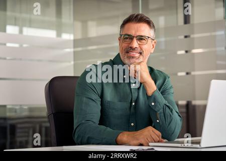 Un uomo d'affari maturo e felice seduto alla scrivania in ufficio che guarda da parte. Foto Stock