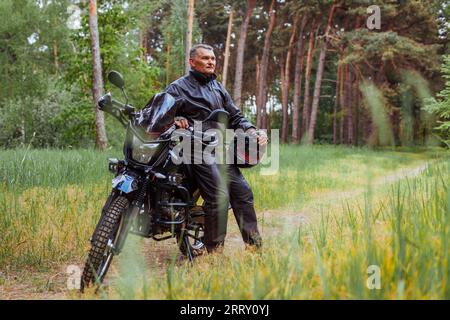Motociclista anziano che si addossa a una moto nella foresta estiva. Uomo in abiti di pelle che si gode la natura tenendo il casco dopo il giro Foto Stock