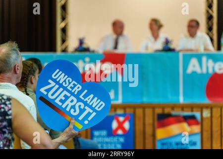 09 settembre 2023, Assia, Gelnhausen: Un sostenitore AfD tiene in piedi un segno a forma di cuore, 'prima il nostro paese! Sullo sfondo, Robert Lambrou (l-r, AfD), presidente dello stato AfD/candidato principale per le elezioni statali, Alice Weidel (AfD), presidente del partito federale, e Tino Chrupalla (AfD), presidente del partito federale, siedono insieme sul podio. L'Hessian AfD tiene la sua campagna d'avvio per le elezioni statali in Assia nella sala dello sport e della cultura di Meerholz. Diverse associazioni e istituzioni manifestano ai margini dell'evento AfD con lo slogan "siamo di più! - Festival per democra Foto Stock