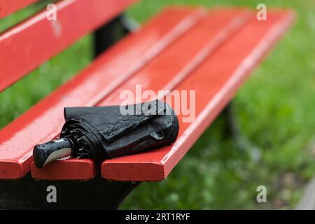 Ombrello nero ripiegato bagnato dimenticato che giace su panca di legno del parco durante la pioggia pesante Foto Stock