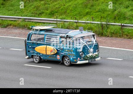 Warren's Vintage VW. Anni '1973 70 settanta Blue Custom Volkswagen Motor Caravan LCV benzina 1584 cc. Bay Window Westfalia camper Van una curiosa auto per matrimoni a noleggio con decorazioni hippie, nastri bianchi e tavola da surf; viaggia a velocità sostenuta sull'autostrada M6 a Greater Manchester, Regno Unito Foto Stock