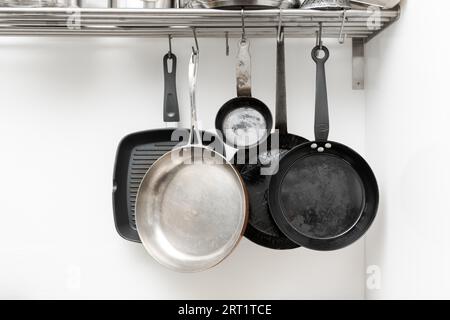 Varie padelle in diverse dimensioni e forme per cucinare e. frittura appesa su ganci in metallo dal ripiano in cucina con parete bianca sullo sfondo Foto Stock