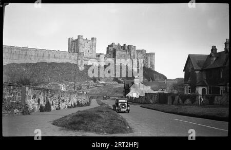 Castello di Bamburgh, Bamburgh, Northumberland, 1940-1953. Vista esterna del castello di Bamburgh, con il castello e la sala dei re sullo sfondo, un monumento commemorativo alla base e le case lungo Front Street in primo piano. Una vista esterna della facciata con il castello e la sala dei re che sporgono da dietro. In primo piano c'è Front Street, con il fronte delle case visibile sul lato destro della strada, e un'auto che guida verso il castello. Alla base della piattaforma rocciosa si trova il castello, dove si trova un memoriale di guerra a forma di croce, che commemora i locali che hanno combattuto in entrambe le guerre mondiali. Foto Stock