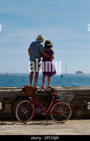 L'uomo tiene la donna intorno alla vita in piedi sulle difese del mare a Portsmouth guardando verso il forte di Spitbank. Due biciclette sotto, appoggiate al muro. Foto Stock