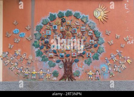 Dettaglio di una decorazione murale in ceramica sulla facciata di un asilo che raffigura un grande albero circondato da bambini, Savona, Liguria, Italia Foto Stock