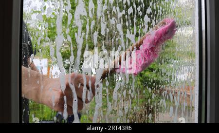 un uomo in piedi sulla strada e visibile attraverso il vetro bagnato lava la finestra di una casa privata con una spazzola rosa, lavando le finestre da un uomo Foto Stock