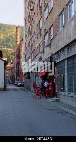 Persone in un caffè di strada con il camino della fabbrica di birra Sarajevska pivara e le lussureggianti colline dietro Sarajevo, Bosnia ed Erzegovina, 10 settembre 2023 Foto Stock