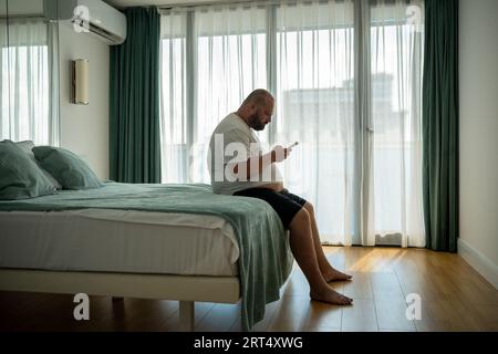 Ragazzo in sovrappeso appeso allo smartphone sul resort seduto nel letto della camera d'albergo in vacanza. Foto Stock