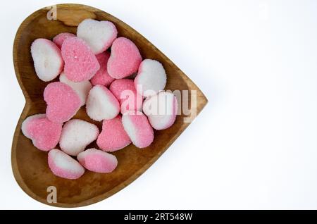 Pila di caramelle di zucchero a forma di cuore su una ciotola di legno a forma di cuore isolata su uno sfondo bianco Foto Stock