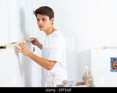 Giovane uomo che controlla la qualità delle piastrelle posate a parete con misuratore di livello Foto Stock