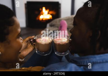 Felice coppia afro-americana che si rilassa a casa davanti al fuoco, facendo un brindisi con cioccolata calda Foto Stock