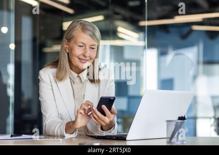 Donna anziana matura con esperienza sul posto di lavoro all'interno dell'ufficio, donna d'affari felice sorridere con il telefono seduto con il computer portatile, donna dai capelli grigi che usa l'app sullo smartphone per scrivere messaggi. Foto Stock