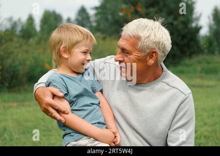 Nonno e nipote si divertono durante una passeggiata nel parco. Buon divertimento per tutta la famiglia. Il nonno del vecchio abbraccia il bambino del ragazzo di 4 anni al giorno di estate. Seni sorridente Foto Stock