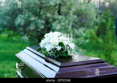 Bara di legno ricoperta da un pesante coperchio con mazzo di fiori bianchi in piedi contro alberi verdi e cespugli sul cimitero prima della sepoltura Foto Stock