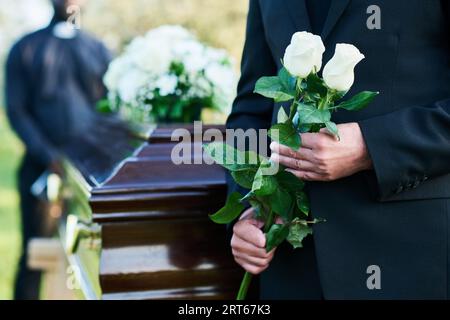 Primo piano di un uomo matue in costume nero con due rose bianche fresche mentre si trova davanti alla macchina fotografica contro la bara con coperchio chiuso Foto Stock