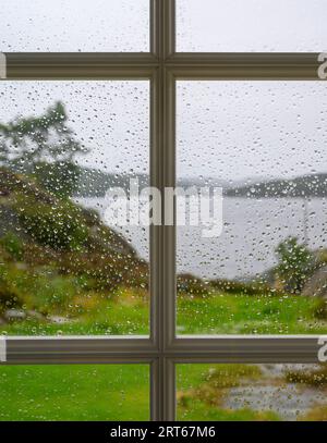 Vista dall'interno verso l'esterno: Vista del paesaggio sfocato (sfocato), del mare e delle isole attraverso una finestra piena di gocce di pioggia a fuoco. Telemark, Norvegia. Foto Stock