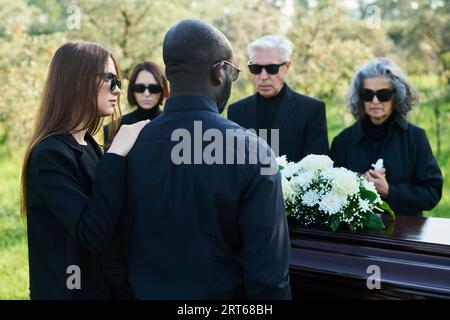 Vista posteriore di una giovane donna in un abbigliamento in lutto che sostiene il marito al funerale, mentre entrambi si trovano davanti alla bara con un mucchio di fiori bianchi Foto Stock