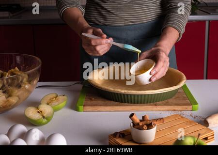 Le mani delle donne spalmano le uova sull'impasto con un pennello da cucina in un piatto da forno, preparazione della torta di mele, preparazione della crostata del Ringraziamento, panetteria autunnale, vite Foto Stock