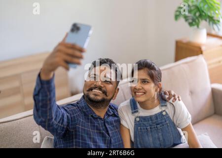 Felice graziosa giovane coppia indiana che si prende selfie a casa seduto sul divano Foto Stock