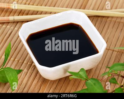 Salsa di soia in un recipiente quadrato bianco su un tappetino di bambù tra coriandolo vietnamita e bacchette Foto Stock