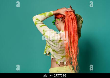 Vista posteriore di una giovane donna elegante con trecce afro arancioni che legano la coda di cavallo mentre si stende su uno sfondo turchese Foto Stock