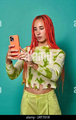 Giovane donna alla moda con lunghi capelli intrecciati che indossa abiti verdi che scatta selfie sul telefono cellulare mentre si trova su uno sfondo turchese Foto Stock