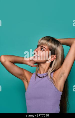 Giovane donna con capelli biondi e piercing che mette le braccia dietro la testa, mostrando l'ascella pelosa su sfondo turchese concetto positivo del corpo Foto Stock