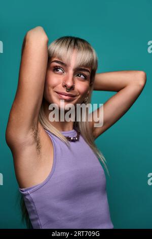 Giovane donna con capelli biondi e piercing che mette le braccia dietro la testa, mostrando l'ascella pelosa su sfondo turchese concetto positivo del corpo Foto Stock