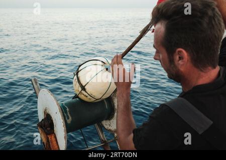 Vista laterale del giovane maschio ispanico che guarda in basso mentre si è in piedi vicino a una ruota a spirale e di giorno si rilascia una boa di sfera bianca nell'acqua di mare di Soller Mallorca Foto Stock