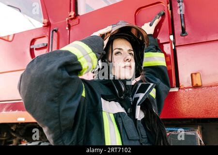 Giovane pompiere senza paura che indossa un casco di protezione regolabile mentre si prepara al lavoro per entrare nel motore antincendio rosso Foto Stock