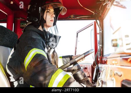 Giovane vigile del fuoco femminile in uniforme protettiva e casco che guida il motore del fuoco rosso mentre lavora al servizio di emergenza guardando lontano Foto Stock