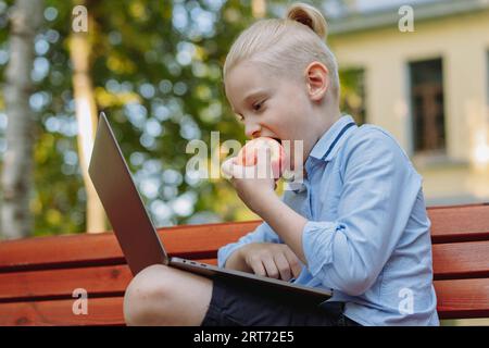 grazioso ragazzo caucasico con coda di cavallo seduto sulla panchina nel parco con un computer portatile che mangia mela. Fare i compiti all'aperto. Immagine con messa a fuoco selettiva Foto Stock