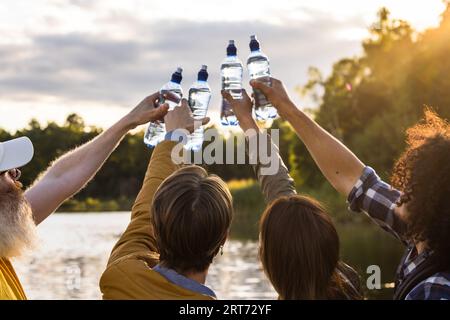 Quattro amici che festeggiano con le bottiglie d'acqua in piedi sulla spiaggia di un lago della foresta durante il bellissimo tramonto e celebrano la vita, foto retroilluminata. Foto di alta qualità Foto Stock