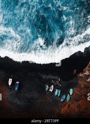 Foto aerea verticale della spiaggia vulcanica di sabbia nera con barche sulla riva e grandi e forti onde nell'oceano - Lanzarote, Isole Canarie. Onde in bl Foto Stock