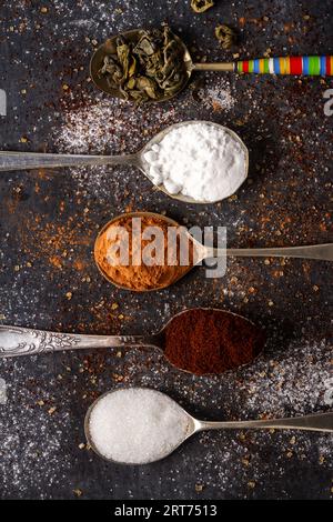 Cucchiai con tè verde, cacao, zucchero a velo e caffè macinato su fondo nero cosparso di zucchero di canna e bianco, caffè e cacao in polvere. Foto Stock