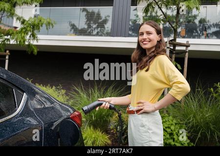 Primo piano di una bella donna che collega il caricabatterie alla sua auto elettrica. Donna progressista che ricarica la sua auto elettrica per strada. Foto Stock