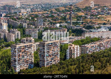Due blocchi di appartamenti in altezza con balconi variamente rivestiti e vetrati si ergono tra le conifere a Tbilisi, in Georgia, sotto il monte Kenisi nel distretto di Nadzaladevi a nord della città. Blocchi prefabbricati, probabilmente dell'era sovietica, si allineano dietro di loro. Un camino a strisce rosse e bianche svetta sopra il complesso. Case indipendenti con tetti colorati coprono i pendii oltre, con un pendio terrazzato in alto a destra. La città ha più di un milione di abitanti. La Cronaca della Georgia, un monumento visibile da lontano, si trova sul monte Kenisi. È stata creata nel 1985 dallo scultore Zurab Tsereteli. Foto Stock