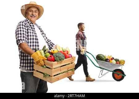 Agricoltore maturo che tiene una cassa con frutta e verdura e un uomo più giovane che spinge una carriola isolata su sfondo bianco Foto Stock