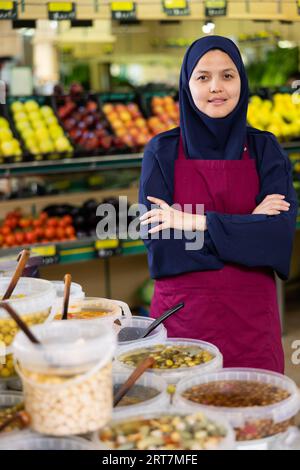 Ritratto di amichevole venditore femminile in hijab e grembiule nella sezione della drogheria del supermercato Foto Stock