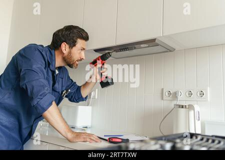 Lavoratore maschio professionista in uniforme utilizza un cacciavite per riparare la cappa di cottura moderna in cucina Foto Stock