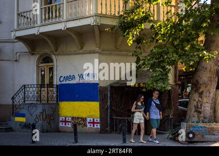 Una bandiera ucraina dipinta sul muro di una casa in una strada di Tbilisi, Georgia, porta le parole "СЛАВА УКРАЇНІ". In inglese: "Gloria all'Ucraina". Sotto ci sono due bandiere georgiane e le parole georgiane rosse "დიდება გმირებს". In inglese: "Gloria agli eroi". Insieme, le due righe formano il saluto ucraino "Gloria all'Ucraina! Gloria agli eroi!". Una coppia cammina mano nella mano davanti all'ingresso di un ristorante. A destra, una cassa di legno contrassegnata "FREE" si trova sotto un albero. Nel 2023, i graffiti contro la Russia e a favore di legami più stretti con l'Europa hanno plasmato il paesaggio urbano di Tbilisi. Il paese stesso Foto Stock